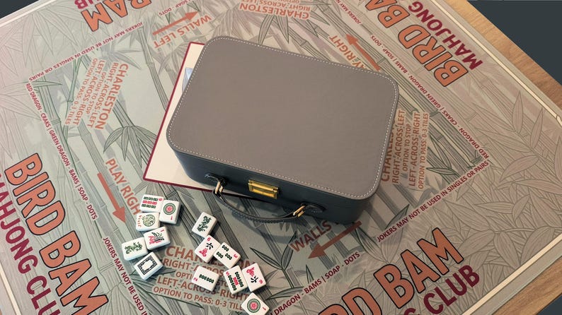 Puede incluir: Un juego de Mahjong gris con un estuche a juego, asa y cierre dorado. Las fichas son blancas con s&iacute;mbolos rojos, verdes y negros. El juego est&aacute; sobre un tablero de juego impreso con el texto "Bird Bam Mahjong Club."