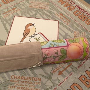 K&ouml;nnte beinhalten: Ein Bird Bam Club Mahjong-Set mit einer Vogelillustration auf einer Karte, einer Pfirsich-Themen-Rolle und einem beigefarbenen Stoffetui. Das Set liegt auf einer Oberfl&auml;che mit Bambusmuster.