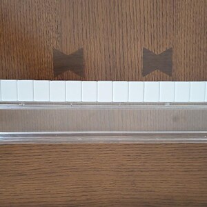 May include: A clear plastic rack designed for holding game tiles, featuring a long, transparent horizontal tray with a hinged front panel and a row of fifteen white rectangular tiles aligned neatly along the top edge of the structure.