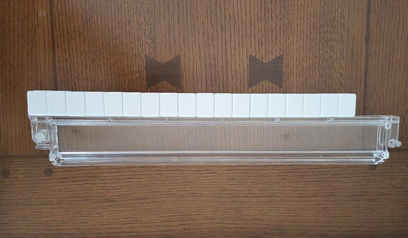 May include: A clear plastic rack designed for holding game tiles, featuring a long, transparent horizontal tray with a hinged front panel and a row of fifteen white rectangular tiles aligned neatly along the top edge of the structure.