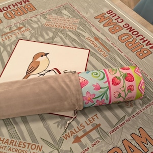 K&ouml;nnte beinhalten: Ein Bird Bam Mahjong Club Spielset. Das Set enth&auml;lt eine Spielmatte mit Bambus und Text, eine Karte mit einer Vogelillustration und eine aufgerollte Matte mit einem Erdbeer- und Blumenmuster. Ein beiger Beutel enth&auml;lt die aufgerollte Matte.