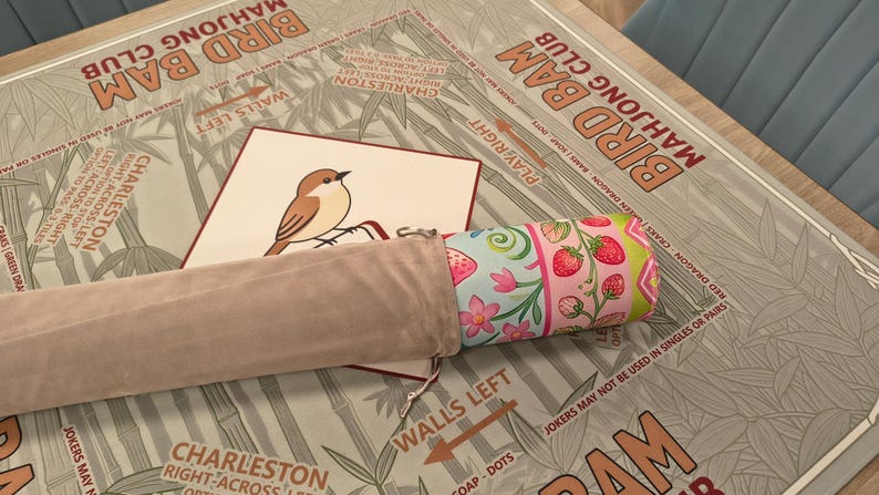 K&ouml;nnte beinhalten: Ein Bird Bam Mahjong Club Spielset. Das Set enth&auml;lt eine Spielmatte mit Bambus und Text, eine Karte mit einer Vogelillustration und eine aufgerollte Matte mit einem Erdbeer- und Blumenmuster. Ein beiger Beutel enth&auml;lt die aufgerollte Matte.