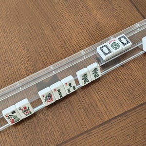 May include: A clear acrylic Mahjong tile rack holding a row of white tiles with green, red, and black symbols. The tiles feature various characters, numbers, and the word JOKER in red or green ink. The rack is a long, narrow, rectangular tray.