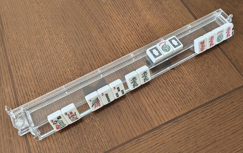 May include: A clear acrylic Mahjong tile rack holding a row of white tiles with green, red, and black symbols. The tiles feature various characters, numbers, and the word JOKER in red or green ink. The rack is a long, narrow, rectangular tray.