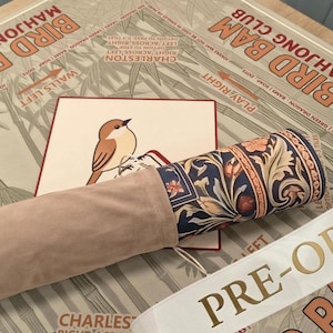 May include: A rolled mat featuring a dark blue floral pattern with orange and cream accents, partially encased in a light beige suede-like sleeve. It rests on a tan surface printed with the text BIRD BAM MAHJONG CLUB and a small bird illustration.