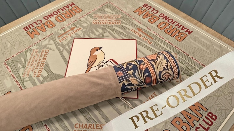 May include: A rolled mat featuring a dark blue floral pattern with orange and cream accents, partially encased in a light beige suede-like sleeve. It rests on a tan surface printed with the text BIRD BAM MAHJONG CLUB and a small bird illustration.