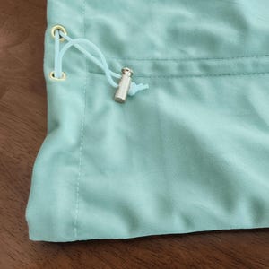 May include: A light mint green fabric pouch featuring a drawstring closure with a matching light mint green cord threaded through two gold-toned metal eyelets, secured by a cylindrical silver-toned metal cord lock at the end of the strings.