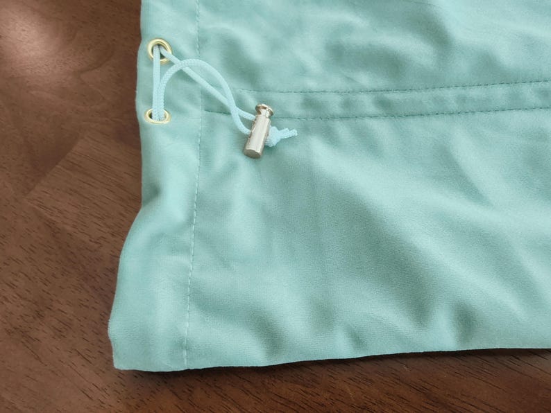 May include: A light mint green fabric pouch featuring a drawstring closure with a matching light mint green cord threaded through two gold-toned metal eyelets, secured by a cylindrical silver-toned metal cord lock at the end of the strings.