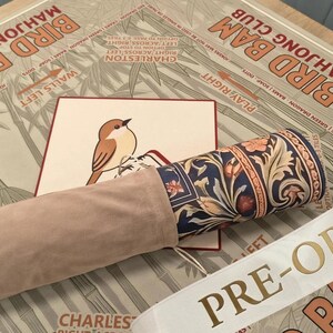May include: A rolled mat featuring a dark blue floral pattern with orange and cream accents, partially encased in a light beige suede-like sleeve. It rests on a tan surface printed with the text BIRD BAM MAHJONG CLUB and a small bird illustration.