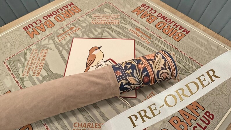 May include: A rolled mat featuring a dark blue floral pattern with orange and cream accents, partially encased in a light beige suede-like sleeve. It rests on a tan surface printed with the text BIRD BAM MAHJONG CLUB and a small bird illustration.