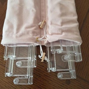 May include: A pale pink velvet drawstring pouch containing several clear acrylic components. The pouch features a white cord with gold-toned metal toggles at the ends. The transparent plastic pieces are rectangular with rounded connectors.