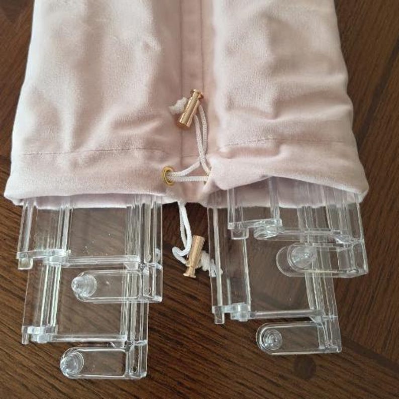 May include: A pale pink velvet drawstring pouch containing several clear acrylic components. The pouch features a white cord with gold-toned metal toggles at the ends. The transparent plastic pieces are rectangular with rounded connectors.