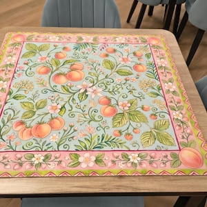 K&ouml;nnte beinhalten: Quadratischer Tisch mit hellem Holzfinish, belegt mit einer dekorativen Matte. Die Matte hat einen hellblauen Hintergrund mit einem Muster aus Pfirsichen, Blumen und gr&uuml;nen Bl&auml;ttern. Der Rand ist rosa und gr&uuml;n.