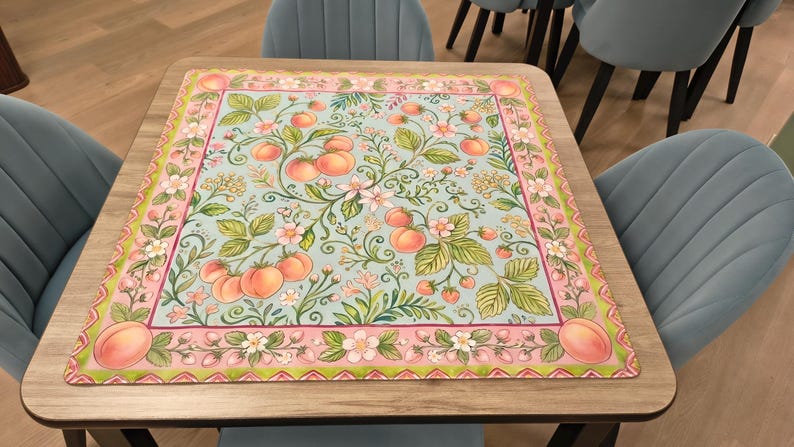 K&ouml;nnte beinhalten: Quadratischer Tisch mit hellem Holzfinish, belegt mit einer dekorativen Matte. Die Matte hat einen hellblauen Hintergrund mit einem Muster aus Pfirsichen, Blumen und gr&uuml;nen Bl&auml;ttern. Der Rand ist rosa und gr&uuml;n.