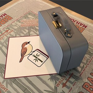 Puede incluir: Un malet&iacute;n de Mahjong azul claro con asa y cierre dorado descansa sobre un tablero de juego. Una tarjeta con una ilustraci&oacute;n de un p&aacute;jaro y el texto "BIRD BAM MAHJONG CLUB" est&aacute; frente al malet&iacute;n.