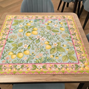 K&ouml;nnte beinhalten: Ein quadratischer Tisch mit einer dekorativen Tischdecke mit einem botanischen Design. Die Tischdecke hat einen hellblauen Hintergrund mit gelben Zitronen, gr&uuml;nen Bl&auml;ttern und wei&szlig;en Bl&uuml;ten. Der Rand ist rosa und gr&uuml;n. Der Tisch ist von blauen St&uuml;hlen umgeben.