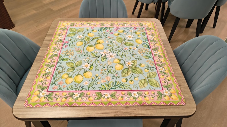 K&ouml;nnte beinhalten: Ein quadratischer Tisch mit einer dekorativen Tischdecke mit einem botanischen Design. Die Tischdecke hat einen hellblauen Hintergrund mit gelben Zitronen, gr&uuml;nen Bl&auml;ttern und wei&szlig;en Bl&uuml;ten. Der Rand ist rosa und gr&uuml;n. Der Tisch ist von blauen St&uuml;hlen umgeben.