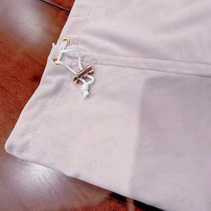 May include: A light pink fabric pouch featuring a white drawstring cord threaded through two gold-toned metal eyelets. The drawstring is secured with a small, cylindrical gold-toned metal toggle, creating a simple closure mechanism on the side.