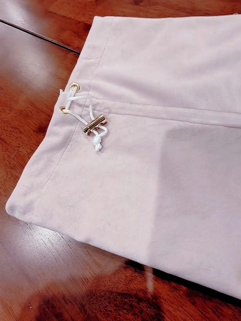 May include: A light pink fabric pouch featuring a white drawstring cord threaded through two gold-toned metal eyelets. The drawstring is secured with a small, cylindrical gold-toned metal toggle, creating a simple closure mechanism on the side.