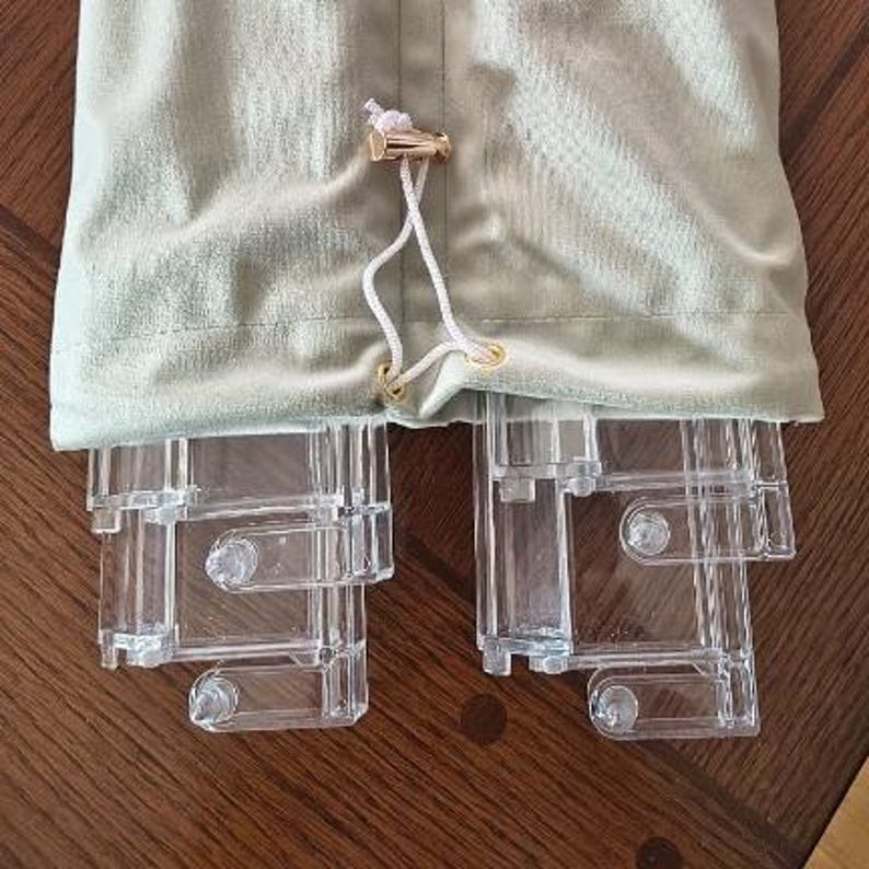 May include: A light sage green velvet drawstring pouch with a metallic gold-toned cord lock, containing two clear acrylic display stands. The stands feature a multi-tiered, rectangular frame design with visible circular connectors and hinges.