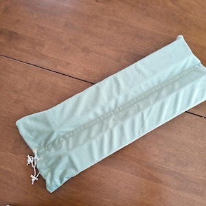 May include: A rectangular, light sage green fabric storage bag featuring a central vertical seam and a white drawstring closure at one end with two small white plastic toggles, designed for holding long items securely within its soft casing.