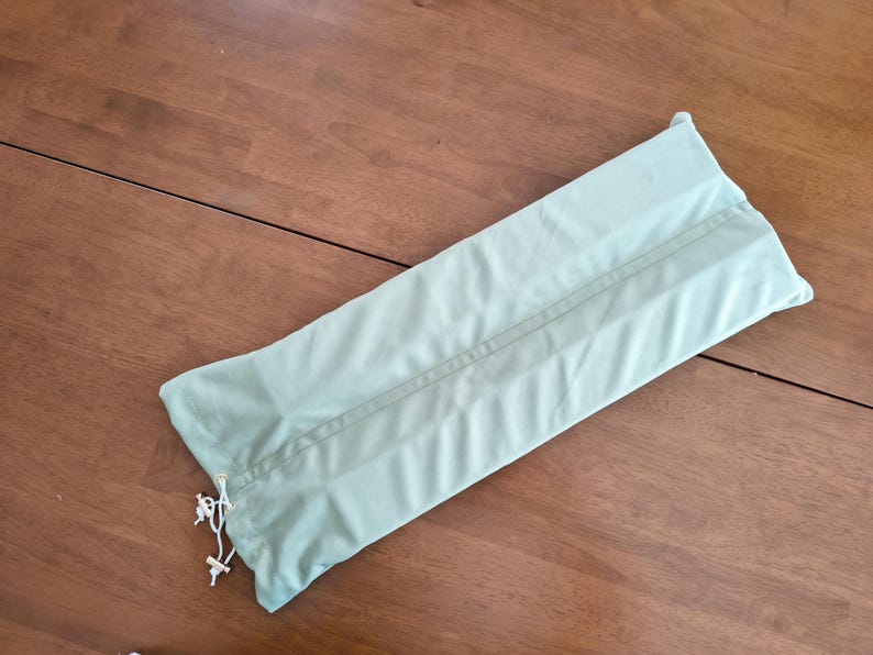 May include: A rectangular, light sage green fabric storage bag featuring a central vertical seam and a white drawstring closure at one end with two small white plastic toggles, designed for holding long items securely within its soft casing.