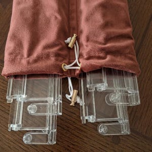 May include: A dusty rose velvet drawstring pouch containing several clear acrylic rectangular components. The pouch features a gold-toned metal toggle closure on white cords, while the transparent plastic pieces are visible protruding from the opening.
