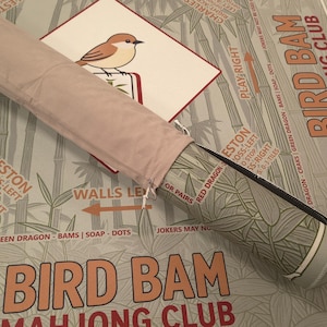 K&ouml;nnte beinhalten: Eine aufgerollte rosa Mahjong-Matte mit passendem Etui. Die Matte zeigt Bambus und den Text "Bird Bam Mahjong Club". Eine Karte mit einer Vogelillustration ist sichtbar. Das Etui hat ein gr&uuml;n-graues Muster.