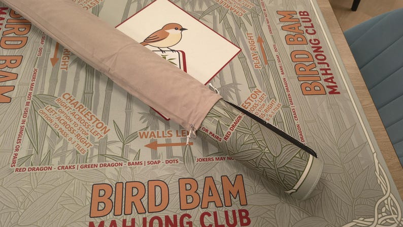 K&ouml;nnte beinhalten: Eine aufgerollte rosa Mahjong-Matte mit passendem Etui. Die Matte zeigt Bambus und den Text "Bird Bam Mahjong Club". Eine Karte mit einer Vogelillustration ist sichtbar. Das Etui hat ein gr&uuml;n-graues Muster.