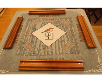May include: A Bird Ram game set with a gray playing surface featuring bamboo illustrations and text. The center has a diamond with a bird and bamboo design. Four wooden tile racks are placed around the board.