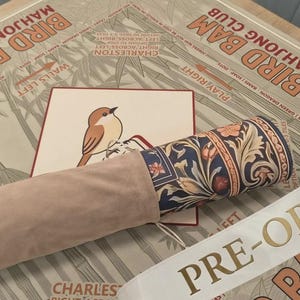 Puede incluir: Una alfombrilla de tela enrollada con un patr&oacute;n floral azul oscuro y detalles en naranja, parcialmente dentro de una funda de ante beige claro. La alfombrilla descansa sobre una superficie color canela con el texto BIRD BAM MAHJONG CLUB.