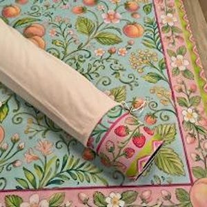 May include: A rolled fabric tube with a light beige exterior, revealing a patterned end featuring red strawberries and green leaves on a pink background. It rests on a flat textile surface decorated with a light blue botanical print of peaches.