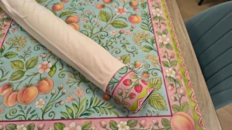 May include: A rolled fabric tube with a light beige exterior, revealing a patterned end featuring red strawberries and green leaves on a pink background. It rests on a flat textile surface decorated with a light blue botanical print of peaches.