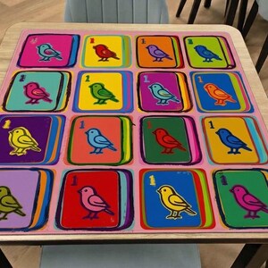 Pode incluir: Um tapete retangular com uma grade de dezasseis quadrados coloridos, cada um exibindo uma ilustra&ccedil;&atilde;o estilizada de um p&aacute;ssaro e o n&uacute;mero um. O design utiliza tons vibrantes de rosa, amarelo, laranja, azul, verde e vermelho, com uma borda rosa claro.