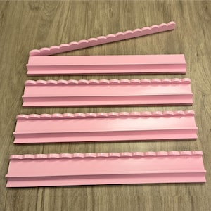 May include: A set of four light pink wall-mounted display shelves, each featuring a decorative scalloped top edge and a flat horizontal ledge for holding items. The shelves are arranged in a parallel formation with one piece placed diagonally.