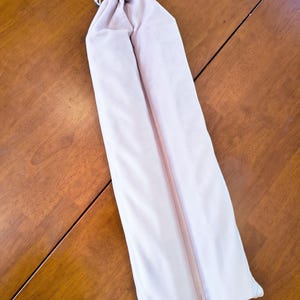 May include: A long, pale pink fabric drawstring pouch with a smooth texture. The bag features a white cord closure at the top, secured with a small, cylindrical wooden bead, designed to hold elongated items in a simple, vertical orientation.