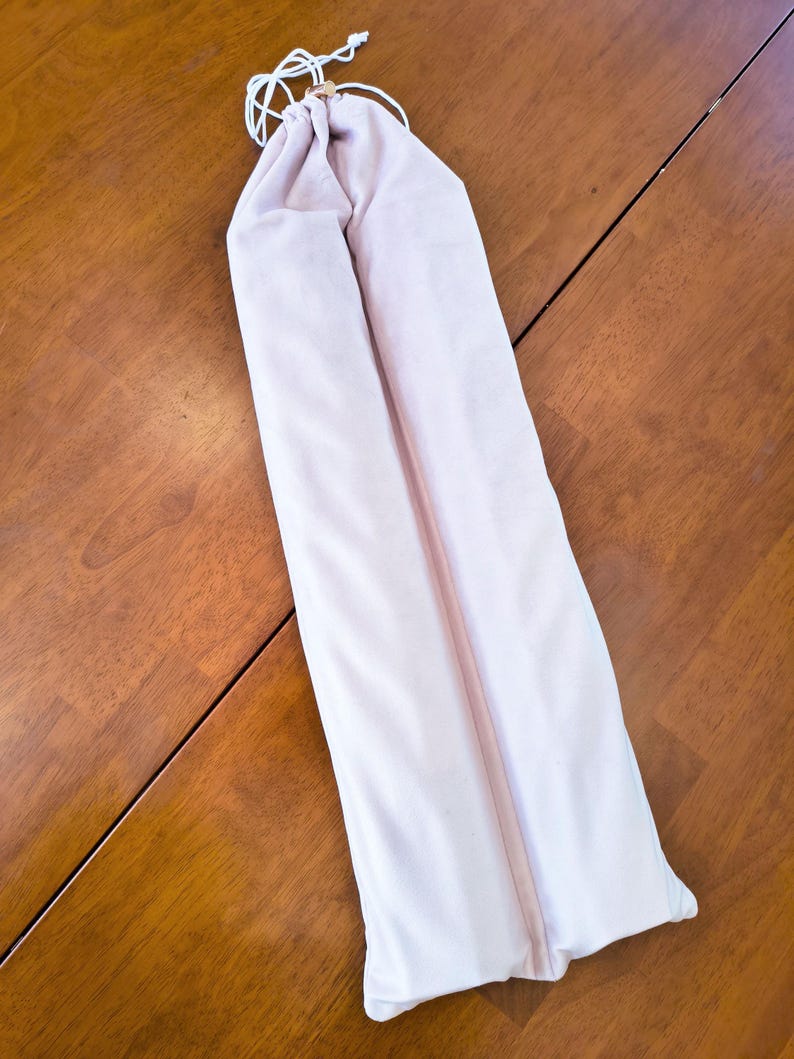May include: A long, pale pink fabric drawstring pouch with a smooth texture. The bag features a white cord closure at the top, secured with a small, cylindrical wooden bead, designed to hold elongated items in a simple, vertical orientation.