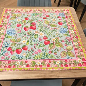 K&ouml;nnte beinhalten: Eine quadratische Tischmatte mit Erdbeermuster. Die Matte hat einen hellblauen Hintergrund mit roten Erdbeeren, gr&uuml;nen Bl&auml;ttern, wei&szlig;en Bl&uuml;ten und rosa und gr&uuml;nen R&auml;ndern. Die Matte liegt auf einem Holztisch.