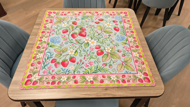 K&ouml;nnte beinhalten: Eine quadratische Tischmatte mit Erdbeermuster. Die Matte hat einen hellblauen Hintergrund mit roten Erdbeeren, gr&uuml;nen Bl&auml;ttern, wei&szlig;en Bl&uuml;ten und rosa und gr&uuml;nen R&auml;ndern. Die Matte liegt auf einem Holztisch.