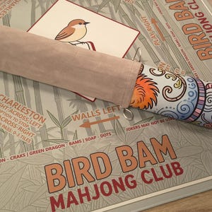 K&ouml;nnte beinhalten: Ein Bird Bam Mahjong Club Spielset. Das Set enth&auml;lt einen farbenfrohen, gemusterten Mahjong-Steinehalter, eine Karte mit einer Vogelillustration und eine Spielmatte mit Bambusmotiv. Der Text "Bird Bam Mahjong Club" ist sichtbar.