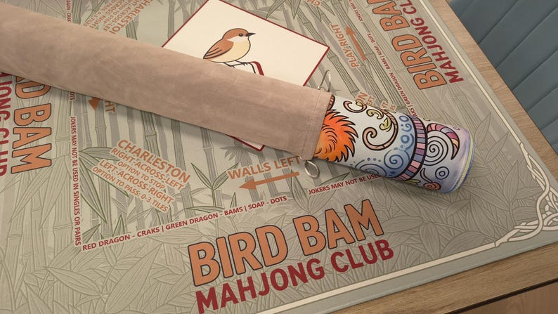 K&ouml;nnte beinhalten: Ein Bird Bam Mahjong Club Spielset. Das Set enth&auml;lt einen farbenfrohen, gemusterten Mahjong-Steinehalter, eine Karte mit einer Vogelillustration und eine Spielmatte mit Bambusmotiv. Der Text "Bird Bam Mahjong Club" ist sichtbar.