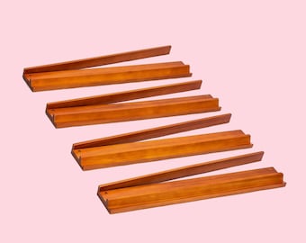 May include: Six wooden tile racks in a warm brown hue are arranged on a light pink background. Each rack has a grooved base to hold tiles and a slightly angled back for visibility. The racks are designed for a board game.