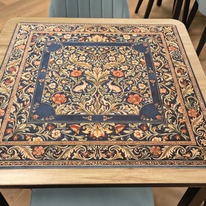 K&ouml;nnte beinhalten: Quadratischer Holztisch mit detailliertem Blumenmuster in Marineblau, Beige und Orange. Der Tisch ist von hellblauen St&uuml;hlen umgeben. Das Design des Tisches weist komplizierte Muster auf.