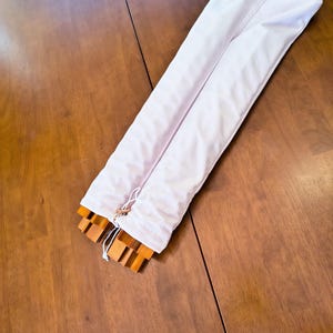 May include: A long, rectangular white fabric storage bag with a drawstring closure at one end. The bag is partially open, revealing several stacked, light brown wooden slats or components inside the protective sleeve.