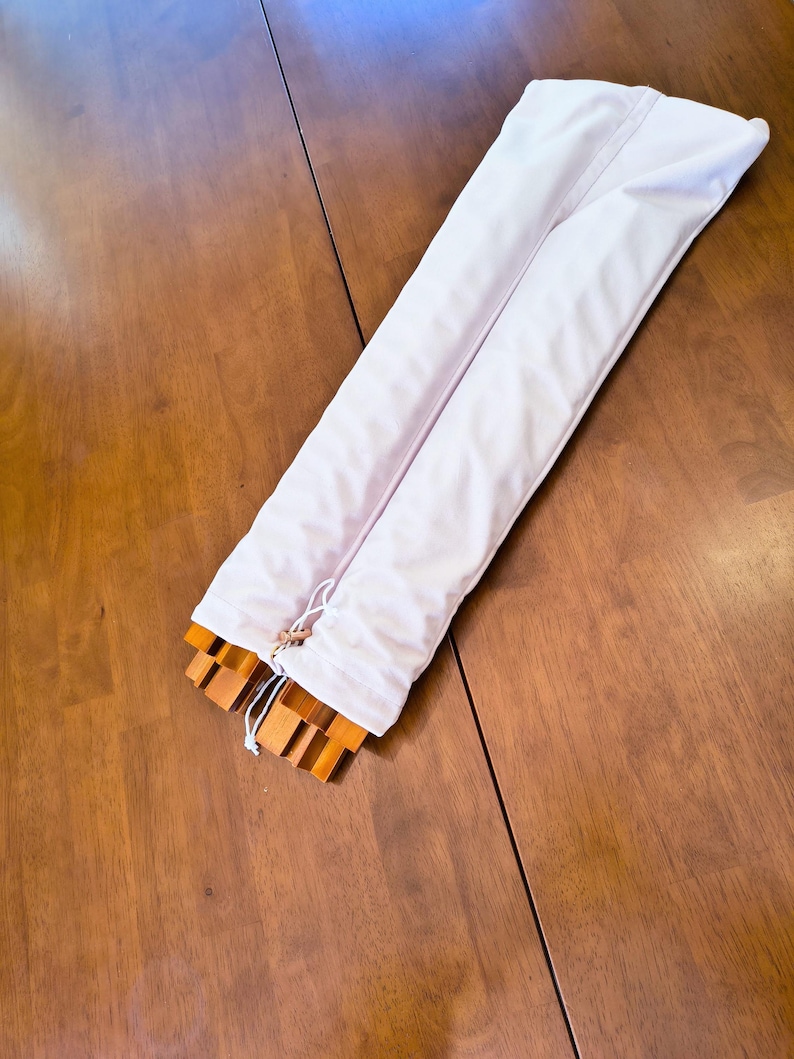 May include: A long, rectangular white fabric storage bag with a drawstring closure at one end. The bag is partially open, revealing several stacked, light brown wooden slats or components inside the protective sleeve.