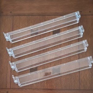 May include: Four clear plastic refrigerator door shelf bins arranged in a parallel formation. Each rectangular bin features a transparent body with integrated side mounting clips and a slightly raised front edge for secure storage of items.