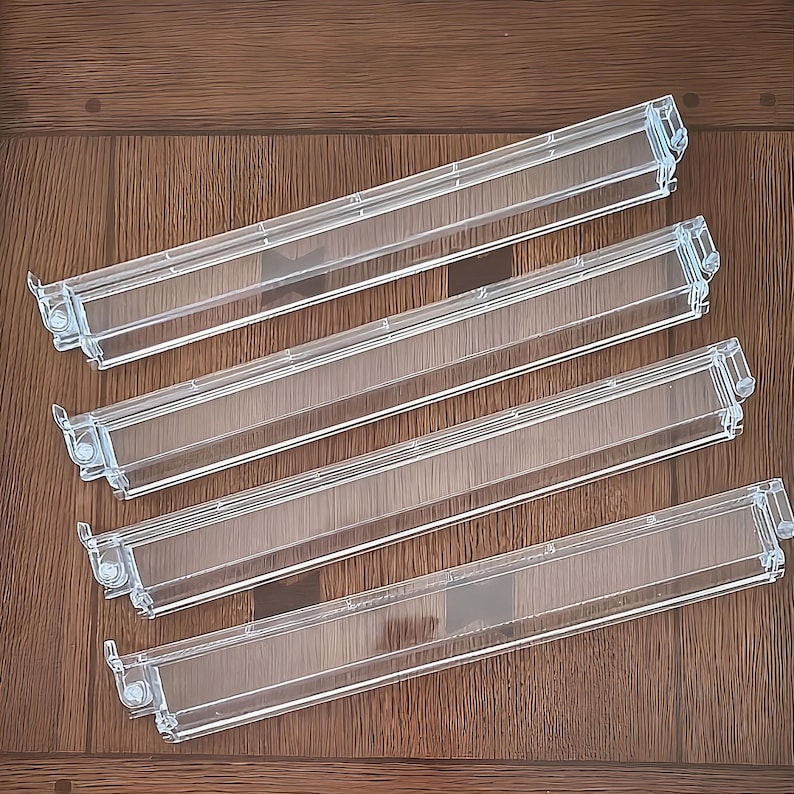 May include: Four clear plastic refrigerator door shelf bins arranged in a parallel formation. Each rectangular bin features a transparent body with integrated side mounting clips and a slightly raised front edge for secure storage of items.