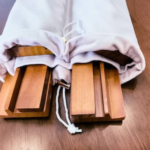May include: Two rectangular wooden stands with grooved surfaces for holding items, partially tucked inside a light-colored fabric drawstring bag. The stands feature a smooth, polished finish and are made of a medium-toned brown wood.