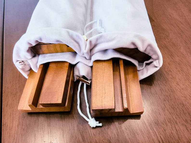 May include: Two rectangular wooden stands with grooved surfaces for holding items, partially tucked inside a light-colored fabric drawstring bag. The stands feature a smooth, polished finish and are made of a medium-toned brown wood.