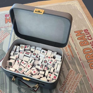 May include: An open gray Mahjong set case with a gold clasp, filled with white tiles featuring red and black symbols. The case sits on a game board with text that reads "Bird Bam Mahjong Club".